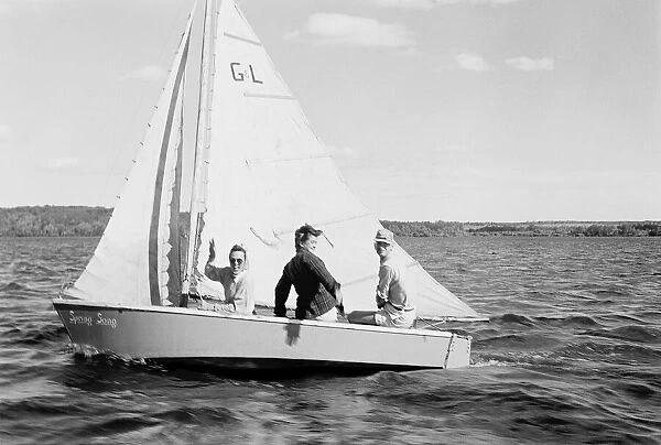 Historical image of three people sailing on Baptiste Lake in a sailboat, the woman waving at the camera, circa 1940; Athabasca County, Alberta, Canada
