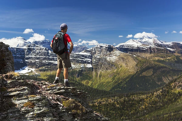 Male Hiker Standing On Top Of Mountain Ridge Overlooking Snow Peaked Mountains And Forest Valley Below With Blue Sky And Clouds; Waterton, Alberta, Canada