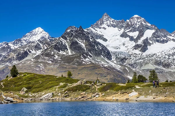 People hiking along the shore of the small mountain lake Grunsee with mountain tops of the Swiss Alps in the background at Zermatt, Switzerland