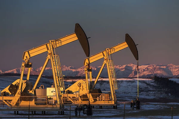 Two Pump Jacks At Sunrise Reflecting The Warm Morning Light In A Snow Covered Field And Snow Covered Foothills And Mountains In The Background Lit By The Morning Sun; Alberta, Canada
