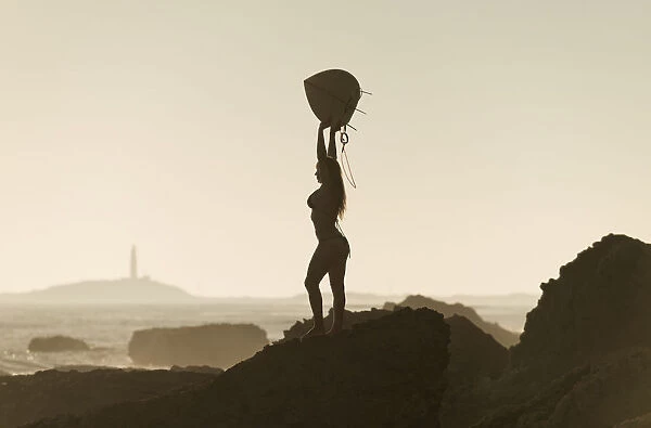 Silhouette of a female surfer holding her surfboard up above her head while standing on a rock at the coast; andalusia spain