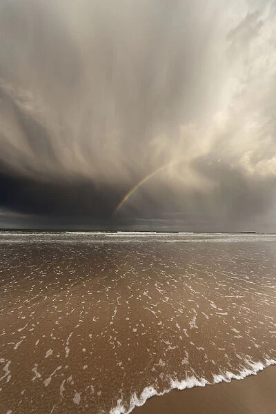 Tide Coming Up Onto The Beach With A Rainbow In The Clouds; Bamburgh, England