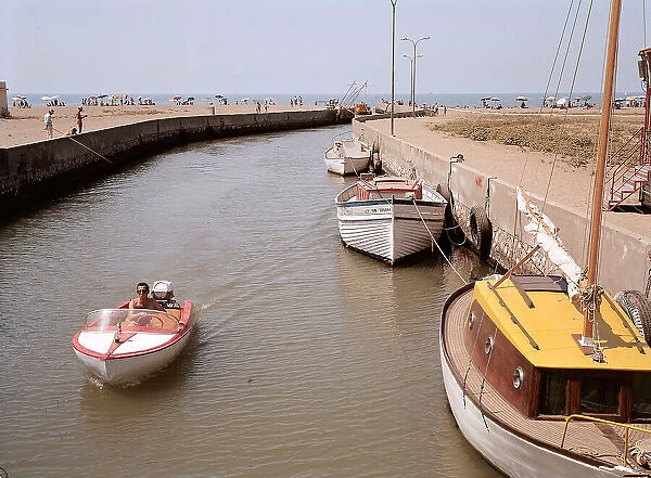 A canal in Marina di Grosseto leading to the sea. A few boats are moored along a bank. In the foreground a motorboat