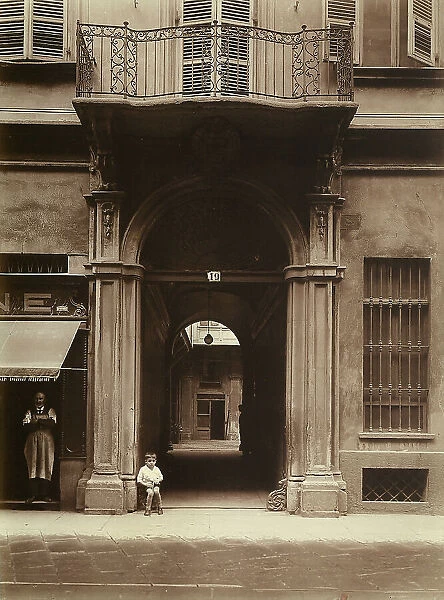 The entrance to an upper-class building in Turin with an elegant portal and a balcony above. A child is sitting on one of the two wrought-iron decorations set near the lower hinges of the door. On the left a man in an apron is standing in the door of a shop