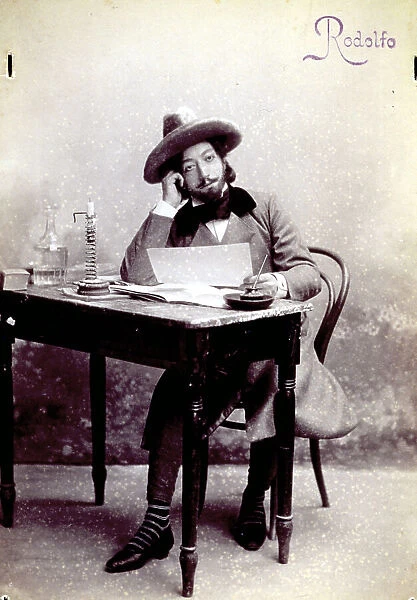 Full-length portrait of a young man playing the part of the poet Rodolfo, in la Boheme by Giacomo Puccini. The man is seated at a table as if he were writing a letter. On the table are an inkpot, a candle holder, a book and a bottle of water