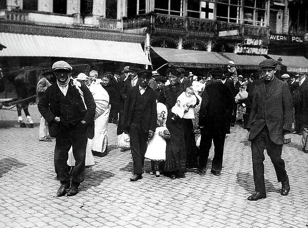 The image portrays a group of refugees, men, women and children, carrying their household goods, in Brussels. In the background, some shops of the city can be seen