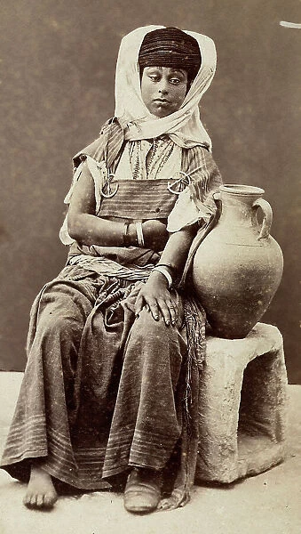 The image shows a lady sit down on the ground, holding a jug for water; she wears a traditional dress covering also her head, forming a turban. The woman comes from a Bedouin tribe of northern Africa