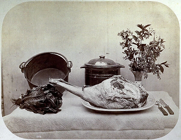 Still life consisting of a pot and a casserole with a vase of flowers in the background. In the foreground a basket of lettuce and an enormous piece of meat on a tray, on the right a fork and knife
