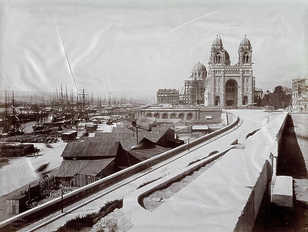The neo-byzantine cathedral known as la Major in Marseilles. Below the church, view of the harbor dotted with ships at anchor. Along the pier freight piled up and freight wagons. In the foreground a road