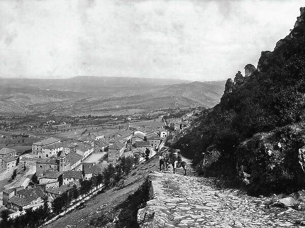 Panoramic view of the town of San Marino. Along the street that leads to the town is a man on a mule chatting with two passersby