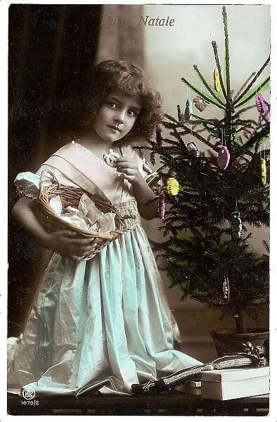 Portrait of a young little girl while eating a cookie and carrying a basket under the Christmas tree, Christmas greeting post-card, with a Buon Natale inscription on the front side and a personal dedication on the back side, the postage stamp indicates the date of 24th of December 1914, Peschiera, Verona, Italy