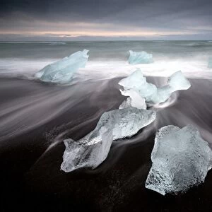 Glassy pieces of ice on volcanic black sand beach with blurred waves, near Jokulsarlon Lagoon