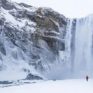 One person in red jacket walking in the snow towards Skogafoss waterfall in winter
