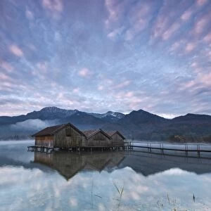 Pink clouds at sunset and wooden huts are reflected in the clear water of Kochelsee
