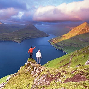 Two hikers on top of a rock admiring the fjord of Funningur at sunrise, Eysturoy island, Faroe islands, Denmark (MR)
