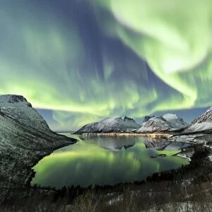 Northern lights and stars light up the snowy peaks reflected in the cold sea Bergsbotn
