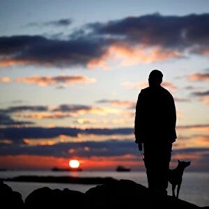 A man watches the sunset with his dog on a breakwater, along the shore of the