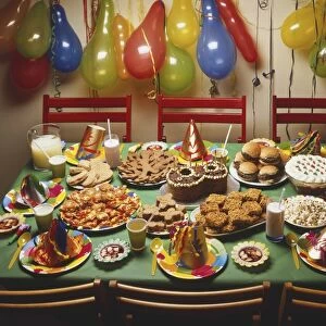 Childrens party display with balloons and chains hanging from wall, table surrounded by six chairs in centre, laid out with cakes, biscuits, puddings, hamburgers and pizza slices, drinks and funny hats