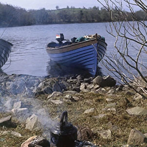 Ireland, brown trout grilling over fire next to fishing rod, small fishing boat moored nearby