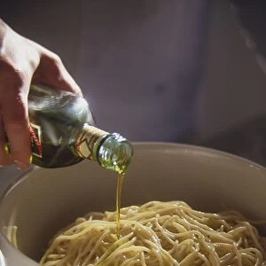 Olive oil being poured from bottle over cooked spaghetti in white bowl