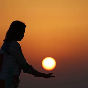 Silhouette of young woman standing by the seashore holding the sun over orange sky