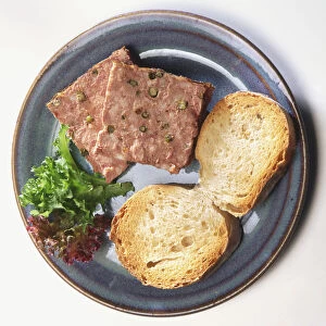 Two square slices of port-flavoured pate seasoned with green peppercorns, served on blue plate with two slices of crusty white bread and salad garnish, view from above