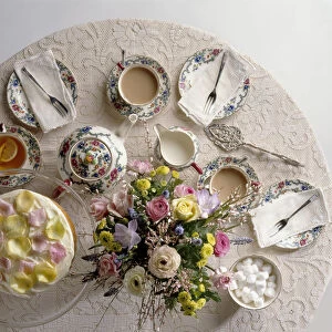 Table set for afternoon tea with bone china tea set, flower arrangement in centre, and cake decorated with rose petals