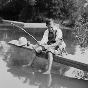 Boy fishing with homemade pole, feet dangling in water, dog sitting by his side