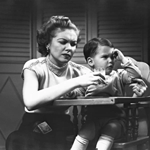 Mother giving glass of milk to son (2-3) sitting in high chair, (B&W)
