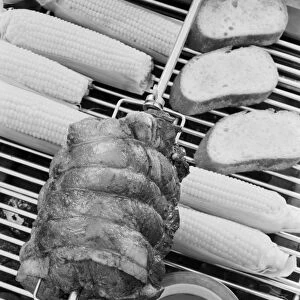 Spit roasting of ham on barbecue grill with corn cobs, close-up