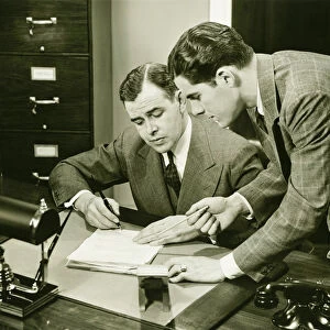 Two young businessmen talking at small desk, (B&W)