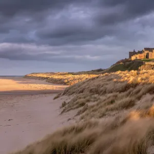 Bamburgh Castle and sand dunes in warm, late evening light with stormy evening sky