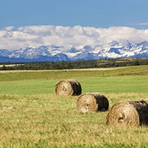 Three Hay Bales In A Field With Mountains In The Background Slightly Snow Covered And Cloudy With Blue Sky; Alberta, Canada