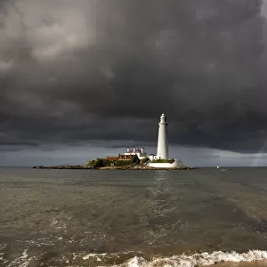 White Lighthouse Illuminated By Sunlight Under A Dark Stormy Sky With A Rainbow; St. Marys Island Northumberland England