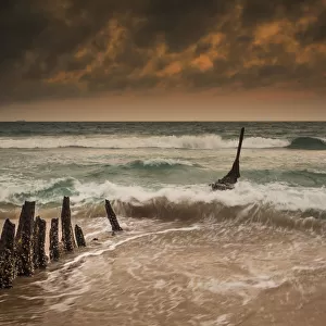 Wooden Posts On A Beach With A Boat Being Tossed In The Water And Waves Crashing Into The Sand Under A Cloudy Sky; Queensland, Australia