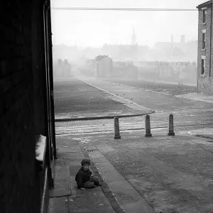 A photograph of a child sitting on the pavement in a Liverpool slum housing area