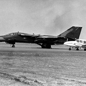 A USAF General Dynamics F-111 Aardvark sits on an airfield next to twin engined civil