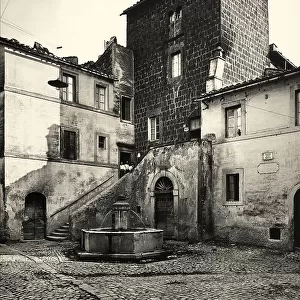 Piazza della Rocca, in Sutri. In the middle of the square there is a fountain with a hexagonal basin. On the right, there is a house from the Medieval period with a mullioned window with two lights