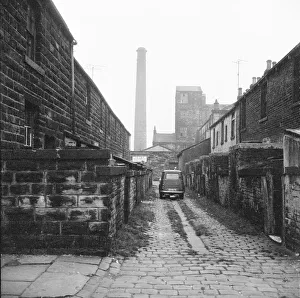 <b class=qtipBold><figcaption>Burnley DES01_01_0538</figcaption></b><br>A car parked in the cobbled ginnel (alley) running between Milton Street and Tentre Street viewed from Richard Street, Burnley, Lancashire, with Pentridge Mill and mill chimney partially visible in the background. The terraces shown here were demolished in the late 1960s. Holly House flats now occupies the site.. Photographed by Eileen Deste between 1966 and 1974.<br><em>© Historic England Archive</em> Burnley DES01_01_0538